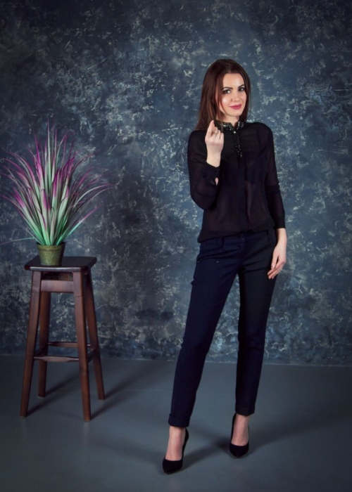 Beautiful young brunette woman, wearing black blouse and dark blue pants, standing in a room with gray walls and houseplant with colorful leaves on a wooden chair
